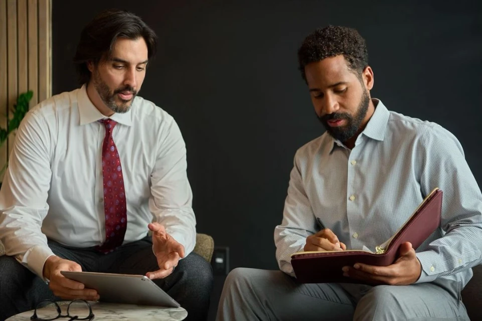 Two business professionals reviewing a tablet and taking notes in a modern office setting, dressed in button-down shirts and ties, indoors.